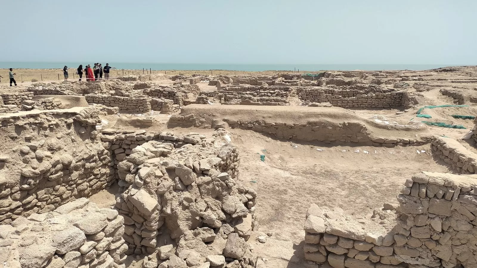 جدران حجرية منخفضة لمدينة أثرية مكشوفة في جزيرة فيلكا بالكويت، والبحر في الخلفية وزوار يقفون عند حافة الحفريات