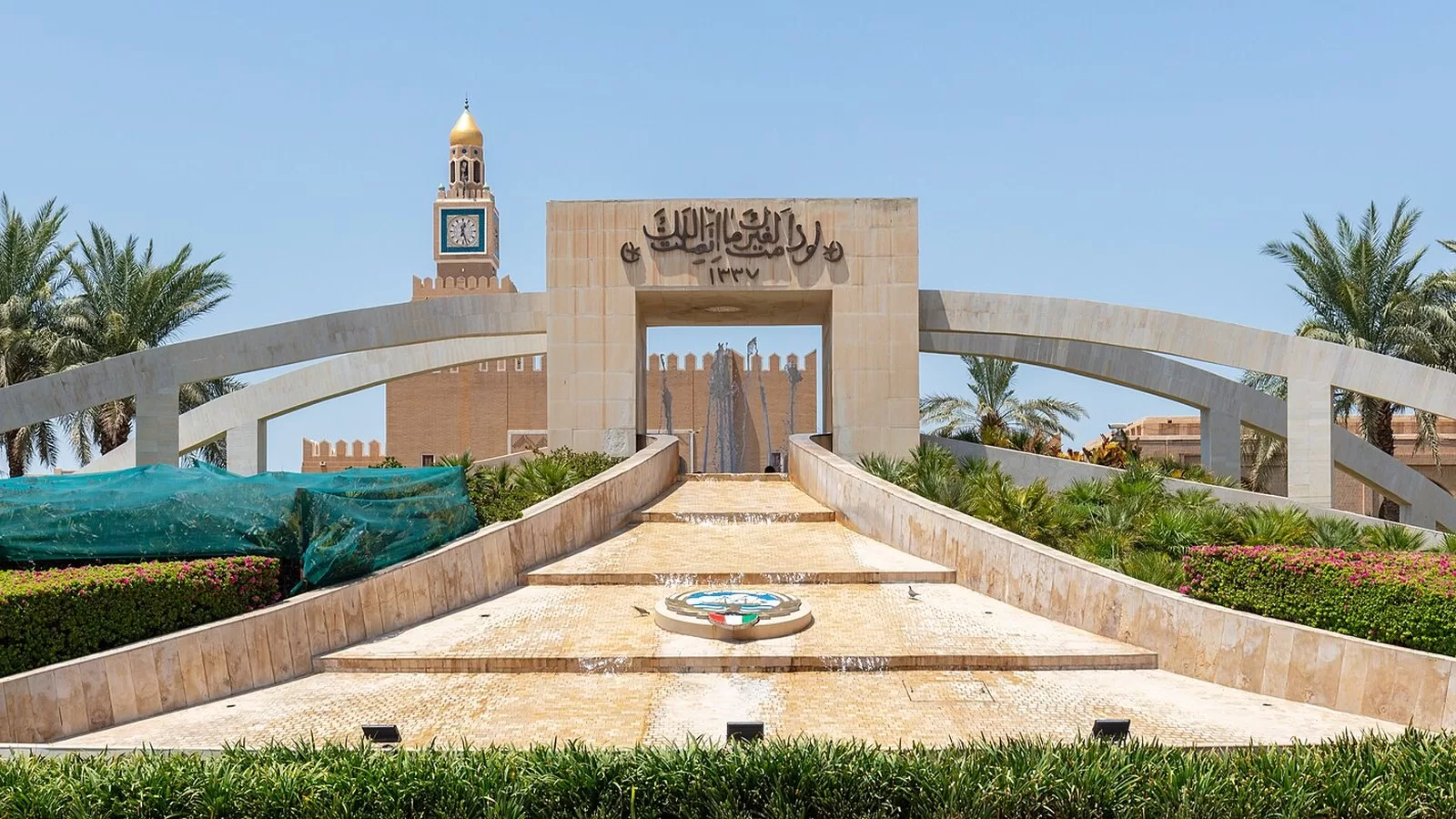 بوابة حجرية ونافورة في ساحة السيف بمدينة الكويت، وخلفها برج ساعة قصر السيف بقبته الذهبية والنخيل على الجانبين