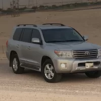سيارة دفع رباعي فضية واقفة على جانب طريق في الجهراء، وخلفها ارض رملية وشاحنة بعيدة