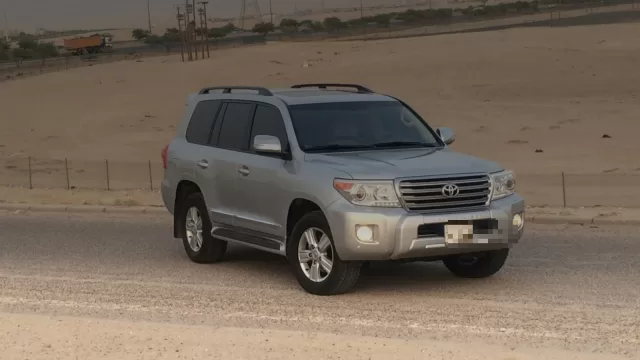 سيارة دفع رباعي فضية واقفة على جانب طريق في الجهراء، وخلفها ارض رملية وشاحنة بعيدة