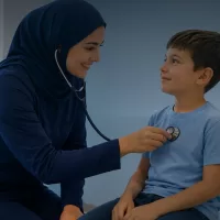ممرضة ترتدي حجابا كحليا تفحص صبيا بسماعة طبية داخل عيادة، والصبي جالس على كرسي ينظر اليها مبتسما