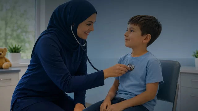 ممرضة ترتدي حجابا كحليا تفحص صبيا بسماعة طبية داخل عيادة، والصبي جالس على كرسي ينظر اليها مبتسما