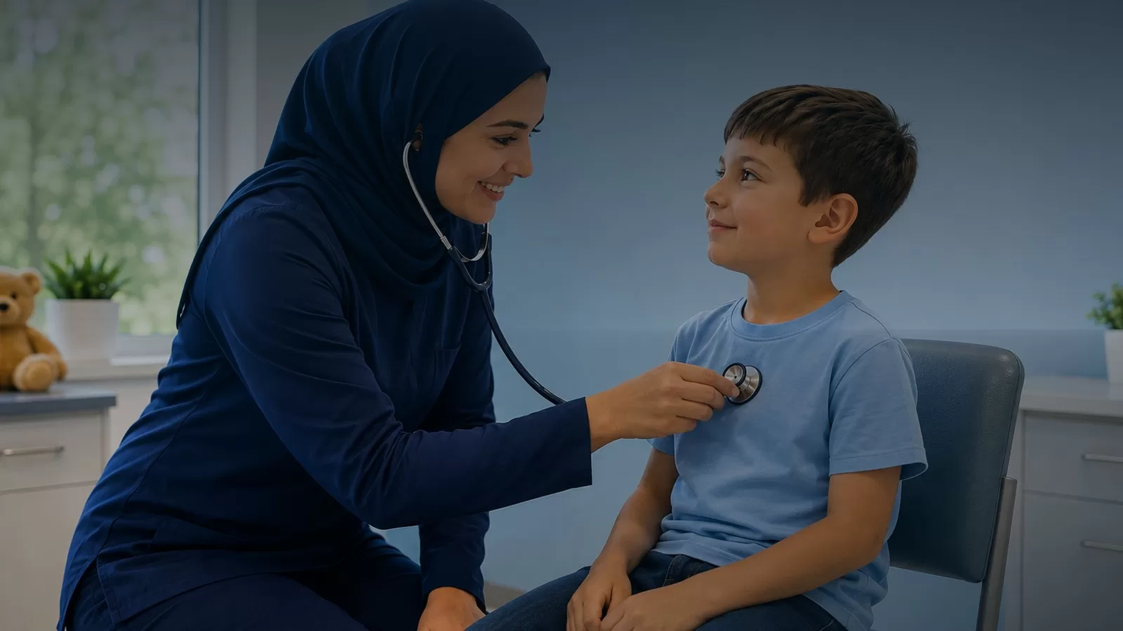 ممرضة ترتدي حجابا كحليا تفحص صبيا بسماعة طبية داخل عيادة، والصبي جالس على كرسي ينظر اليها مبتسما