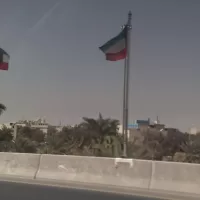 علما الكويت يرفرفان فوق جسر الدائري الرابع، وتحتهما نخيل ومبان سكنية وطريق سريع