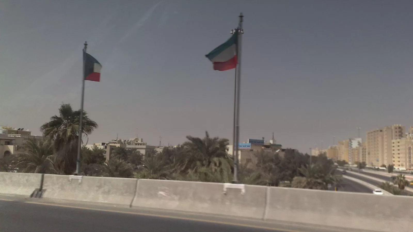 علما الكويت يرفرفان فوق جسر الدائري الرابع، وتحتهما نخيل ومبان سكنية وطريق سريع