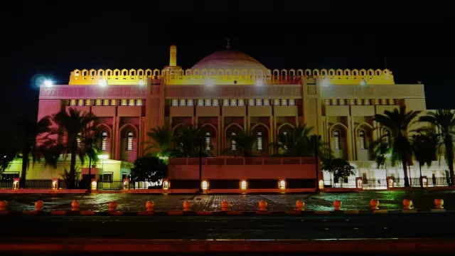 واجهة المسجد الكبير في مدينة الكويت ليلا مضاءة باللون الذهبي، امامها صف من النخيل وسور المسجد، والسماء سوداء فوقها.