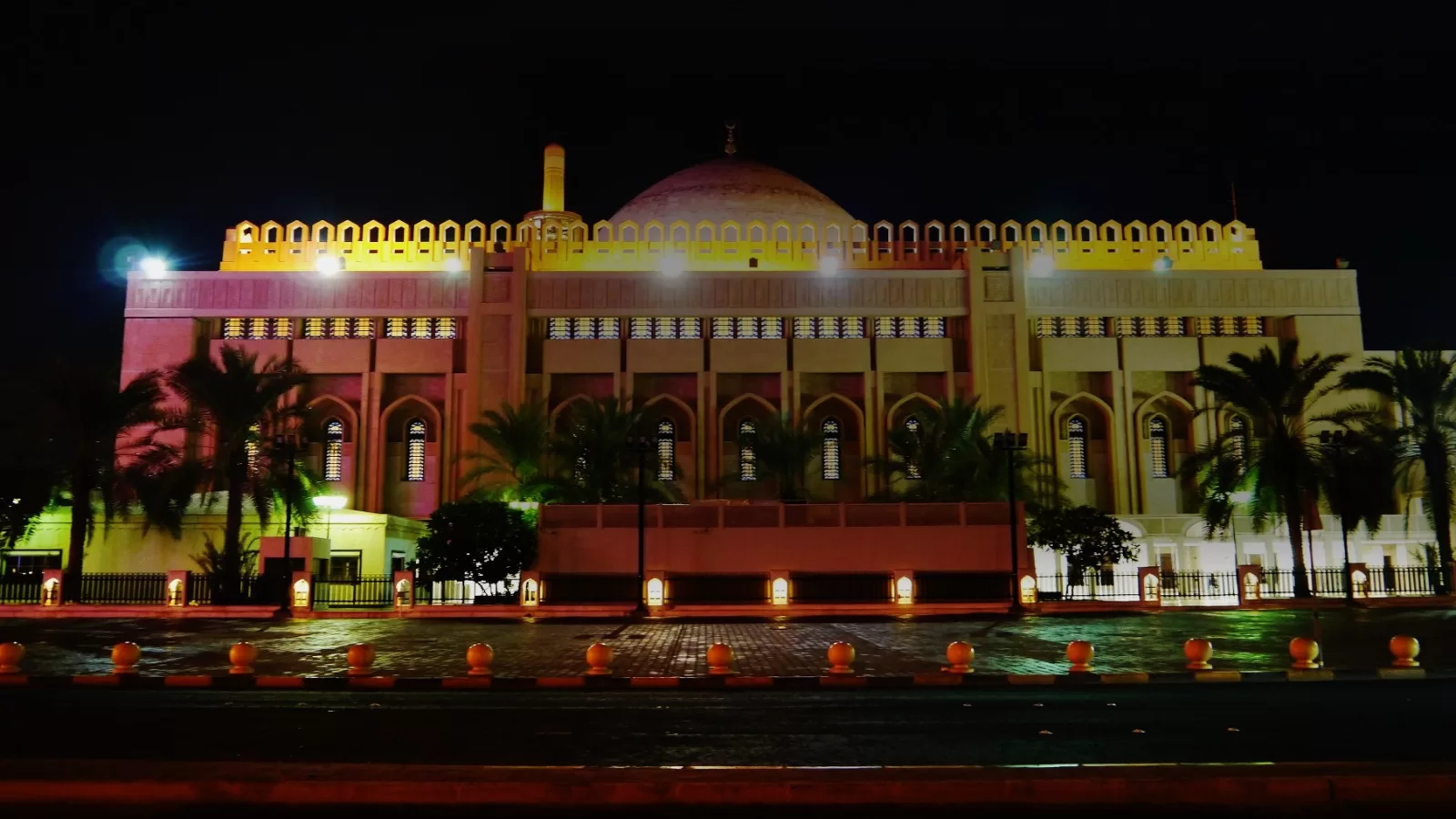 واجهة المسجد الكبير في مدينة الكويت ليلا مضاءة باللون الذهبي، امامها صف من النخيل وسور المسجد، والسماء سوداء فوقها.