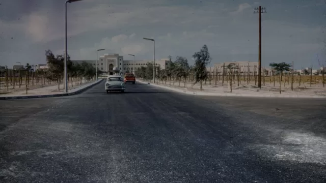 طريق مسفلت في الكويت عام 1961 وعليه سيارتان من طراز الخمسينات، وفي نهايته مبنى حكومي ابيض واسع