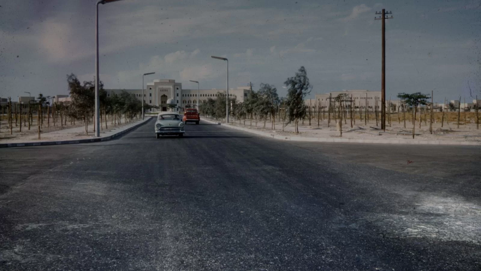 طريق مسفلت في الكويت عام 1961 وعليه سيارتان من طراز الخمسينات، وفي نهايته مبنى حكومي ابيض واسع