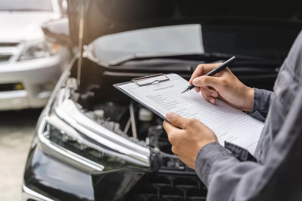 An inspector filling in a checklist in front of a car with its bonnet open